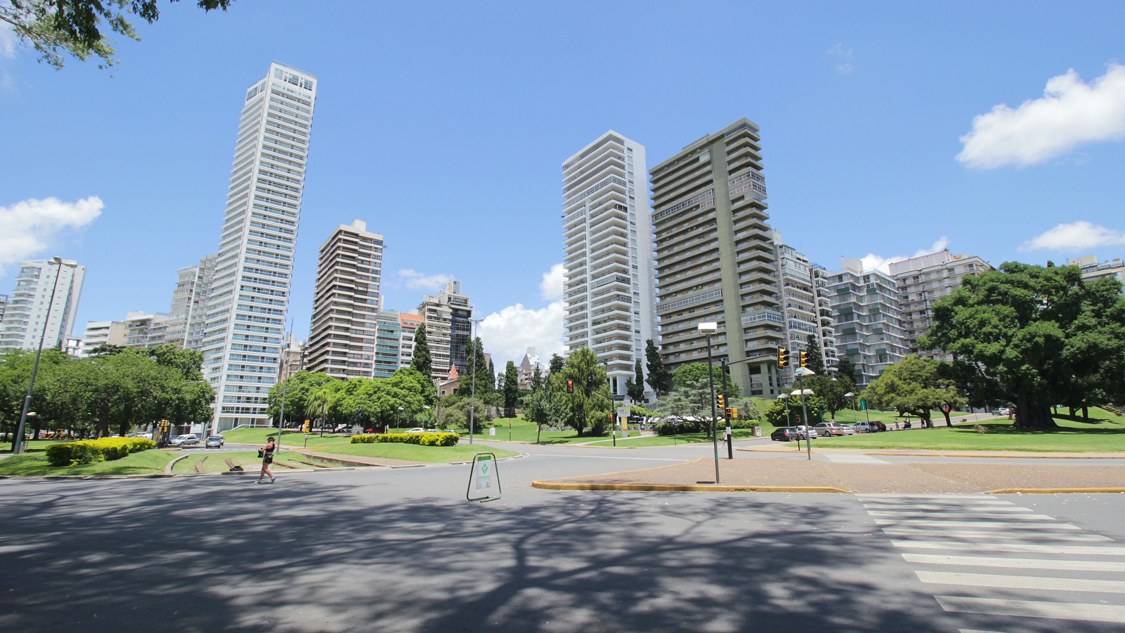 The image captures a portion of the park, featuring preserved railway fragments. A woman is seen crossing the street in the foreground. The vibrant blue sky contrasts with the lush greenery of the trees, creating a picturesque view of the city.